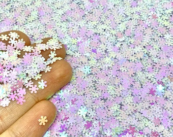 May include: A close-up of iridescent white snowflake glitter. The glitter is scattered on a surface and a hand is holding some of the glitter.
