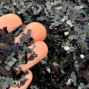 May include: A collection of small, black cat-shaped sequins featuring a holographic finish that reflects various colors. These tiny, flat, cat-shaped pieces are scattered in a pile, with some resting on a surface and others held in a hand.