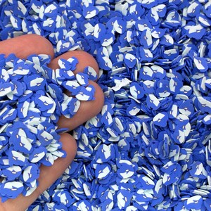 May include: A close-up of blue and white polymer clay whale-shaped sprinkles. The sprinkles are scattered across the image, with some held in a hand. The whale shapes have a blue body and a white belly.