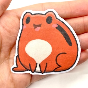 May include: A red frog sticker with a white belly and black eyes. The frog is smiling with its mouth open.