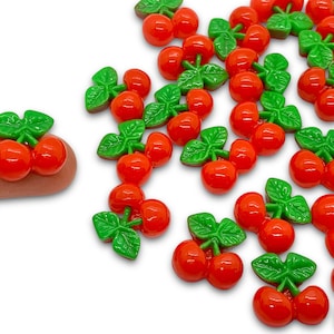 May include: A collection of red and green cherry-shaped embellishments. Each cherry has a green stem and leaves. The cherries are made of plastic and are about 1 inch in diameter.