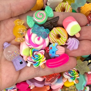 May include: A collection of miniature resin cabochons shaped like various sweets, including lollipops, donuts, ice cream bars, cupcakes, gumball machines, and candies in vibrant colors like pink, green, yellow, and purple, held in an open palm.