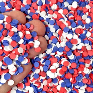 May include: A handful of red, white, and blue confetti. The confetti is in the shape of small, flat circles. The colors are vibrant and the confetti is scattered across the image.