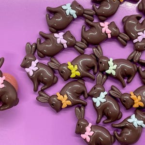 May include: A collection of brown bunny-shaped embellishments with colorful bow accents. The bunnies are arranged on a purple surface. One bunny is held by a finger. These decorative elements are suitable for crafts and Easter-themed projects.
