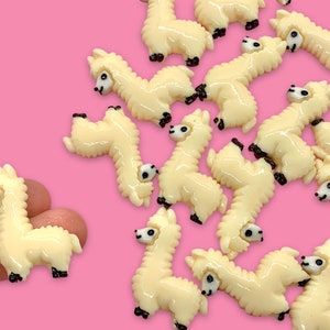 May include: A collection of cream-colored llama charms with black eyes and noses. The charms are arranged on a pink background.