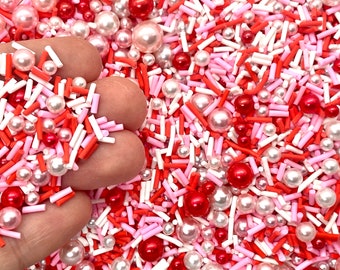 May include: A mix of red, pink, and white sprinkles and pearl-like sprinkles. The sprinkles are in a variety of shapes and sizes.