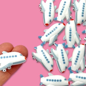 May include: A collection of white and blue airplane charms with red accents. The charms are small and perfect for adding a touch of whimsy to crafts and jewelry.