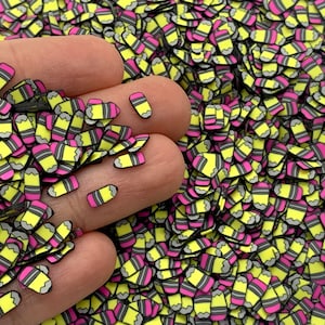 May include: A large quantity of yellow and pink pencil-shaped resin embellishments. The embellishments are scattered on a surface and a hand is holding some of them.