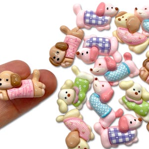 May include: A collection of colorful, miniature dog figurines. The dogs are in various poses and wear outfits in shades of pink, blue, green, and brown. Some have checkered patterns. The figurines are made of a glossy material.