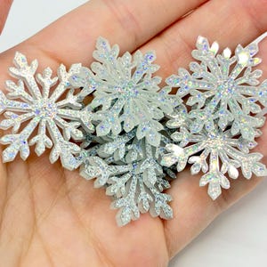May include: A collection of small, white, six-pointed snowflake ornaments featuring a reflective, iridescent finish that catches the light. The snowflakes have intricate, branching arms and are layered together in a cluster.