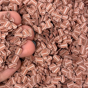 May include: A large collection of small, flat, brown football-shaped embellishments. Each piece features a white outline and white stitched details resembling the laces of a football. The items are uniform in size and shape, creating a dense, textured pile of sports-themed decorative accents for crafting projects.