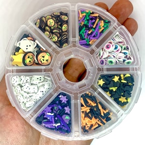 May include: A clear plastic container with eight compartments filled with various Halloween-themed sprinkles. The sprinkles are in different colors, including black, orange, green, purple, and white. The sprinkles are shaped like stars, eyes, pumpkins, ghosts, and other Halloween-themed shapes.
