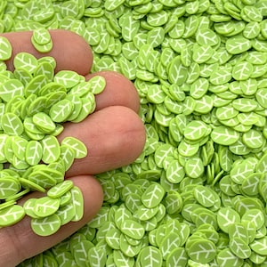 May include: A large quantity of green leaf-shaped polymer clay sprinkles with white outlines. The sprinkles are scattered on a surface and a hand is holding a handful of the sprinkles.