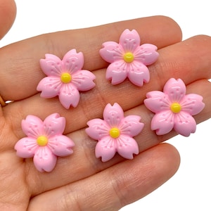 May include: Five pink resin flower embellishments with yellow centers. Each flower has five petals and a textured surface.