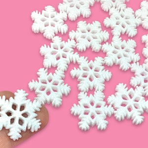 May include: A scattering of white glitter snowflake ornaments on a pink background. The snowflakes are all the same size and shape, and they are arranged in a random pattern.