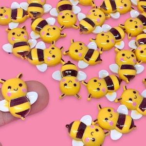May include: A collection of yellow and black bee-shaped resin embellishments with white wings and black eyes. The bees are arranged on a pink surface.