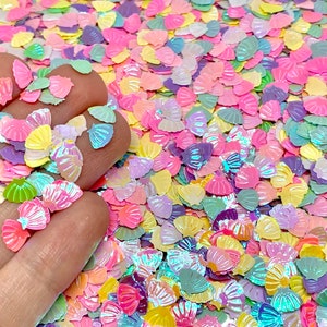 May include: A scattering of iridescent seashell-shaped sequins in pastel colors: pink, yellow, green, and purple. The sequins are small and scattered, creating a shimmering effect. Ideal for crafts, scrapbooking, or embellishments.