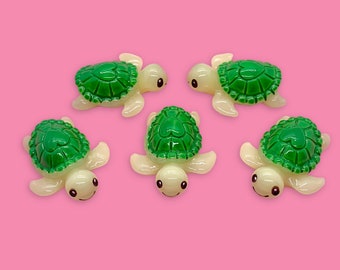 May include: Five small, green and white turtle figurines with smiling faces. The turtles have green shells with a heart design and white bodies.