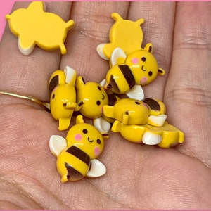 May include: A collection of six yellow and black bee-themed resin charms. The charms feature a variety of designs, including bees with wings, bees with smiling faces, and honeycomb patterns.