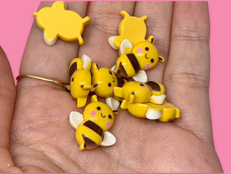 May include: A collection of six yellow and black bee-themed resin charms. The charms feature a variety of designs, including bees with wings, bees with smiling faces, and honeycomb patterns.