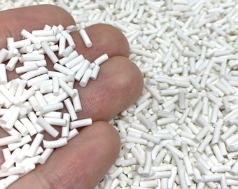 May include: A close-up of a hand holding a pile of small, white, cylindrical beads. The beads are scattered across a surface.