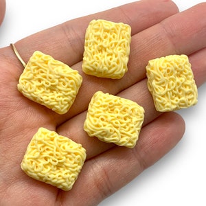 May include: Five yellow plastic miniature instant noodle brick packs. The packs are all the same size and shape, and they have a textured surface that looks like noodles.