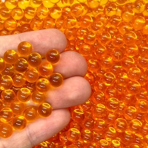 May include: A close-up of a hand holding a cluster of small, translucent orange spheres. The spheres are densely packed, with a glossy, reflective surface. The background is filled with more of the same spheres.