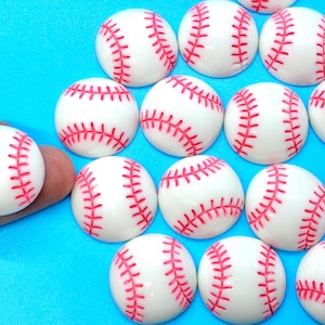 May include: A collection of small, white baseball-shaped cabochons with red stitching. The cabochons are arranged on a bright blue surface. One cabochon is held by a finger. These are likely for crafting or jewelry making.
