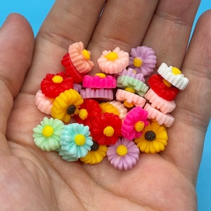 May include: A collection of small, multi-colored flower-shaped cabochons featuring petals in shades of red, orange, yellow, pink, purple, white, and light blue, each with a contrasting yellow or brown center, arranged in a cluster.