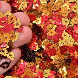 May include: A collection of small, metallic leaf-shaped sequins in shades of red, orange, and gold. Each individual piece features a cutout design in the center, creating a detailed foliage pattern that reflects light with a shiny, reflective finish.