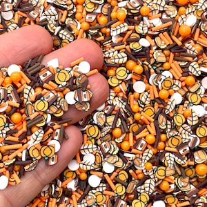 May include: A collection of autumn-themed sprinkles featuring orange, brown, and white colors. The mix includes small shapes of pumpkins, leaves, coffee cups, and acorns, combined with orange and white round sugar pearls and thin rod-shaped pieces.