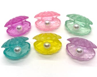 Puede incluir: Seis conchas de plástico iridiscentes con una perla dentro de cada una. Las conchas son de color rosa, verde, amarillo y morado.