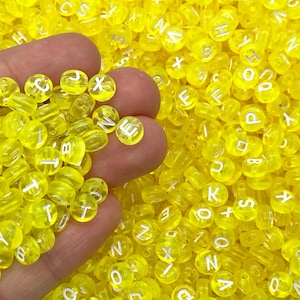 May include: A handful of transparent yellow letter beads with white letters. The beads are round and cylindrical, with a hole through the center. The background is filled with more yellow letter beads. These beads are ideal for jewelry making and crafts.