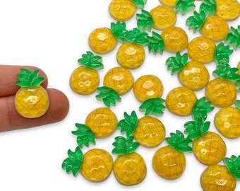 May include: Small, yellow pineapple-shaped embellishments with green leaves. The miniature pineapples have a textured surface and are scattered across a white background. One is held in a person's fingers.