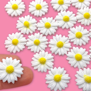 May include: A collection of white daisy-shaped resin embellishments with yellow centers. The daisies are scattered on a pink surface. One daisy is being held by a finger.