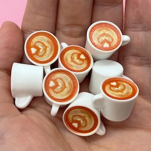 May include: Miniature white coffee cups with latte art. Each cup features a heart-shaped design in the foam, with a warm orange-brown color. The cups have small handles and are arranged in a hand.
