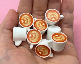 Może przedstawiać: Miniaturowe białe filiżanki do kawy z latte art. Każda filiżanka ma wzór w kształcie serca w piance, w ciepłym pomarańczowo-brązowym kolorze. Filiżanki mają małe uchwyty.