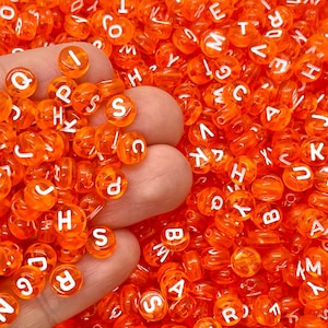 May include: A handful of orange, transparent letter beads with white lettering. The beads are round and scattered, with letters visible. The beads are held in a hand, with many more beads in the background.