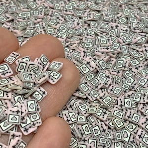 May include: A close-up of a hand holding a pile of small pink and white square-shaped resin charms. Each charm has a black outline and a heart shape with a plus sign inside.