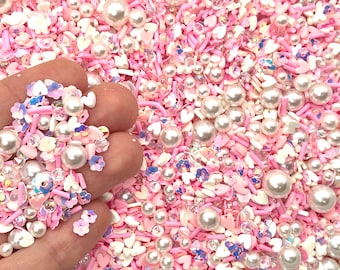 May include: A mix of pink and white sprinkles, including hearts, flowers, and small pearl-like beads.