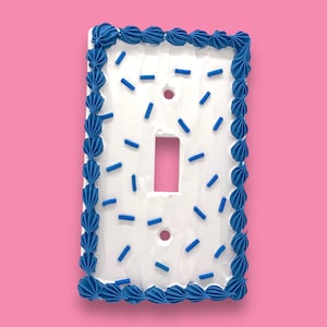 May include: White light switch cover with a blue frosting border and blue sprinkles.