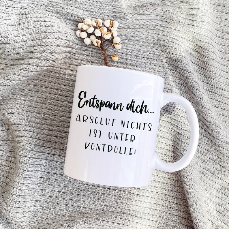 May include: White ceramic mug with the German text "Entspann dich... ABSOLUT NICHTS IST UNTER KONTROLLE!" in black script. The mug has a curved handle and is set against a gray, textured background with a small floral accent.