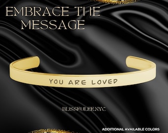 May include: A gold-colored cuff bracelet with the inscription "YOU ARE LOVED." The bracelet is set against a black background with gold accents and the text "EMBRACE THE MESSAGE." Additional colors are available.