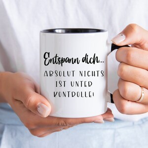 May include: White ceramic mug with a black interior and handle. The mug features the German text "Entspann dich... ABSOLUT NICHTS IST UNTER KONTROLLE!" in black script. The mug is held by a person.