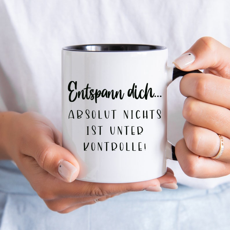 May include: White ceramic mug with a black interior and handle. The mug features the German text "Entspann dich... ABSOLUT NICHTS IST UNTER KONTROLLE!" in black script. The mug is held by a person.