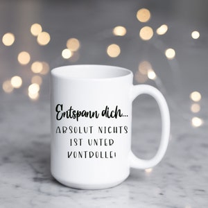 May include: White ceramic mug with a large handle. The mug features black text that reads "Entspann dich... ABSOLUT NICHTS IST UNTER KONTROLLE!" The background is blurred with bokeh lights.
