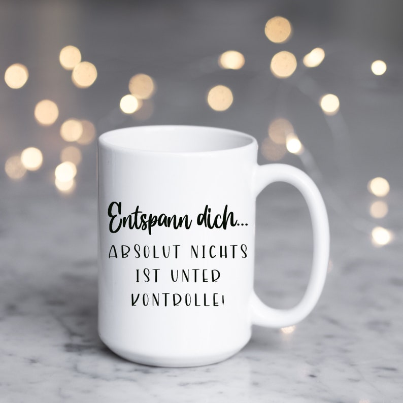 May include: White ceramic mug with a large handle. The mug features black text that reads "Entspann dich... ABSOLUT NICHTS IST UNTER KONTROLLE!" The background is blurred with bokeh lights.