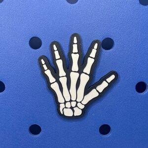 May include: A white skeleton hand charm with black outlines on a blue background.