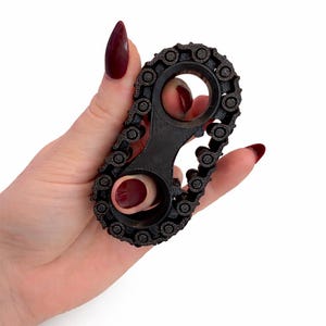 May include: A black, chain-link style fidget toy with a central handle and two circular openings. The toy is held in a hand with burgundy nail polish, set against a white background. The chain links have a textured appearance.