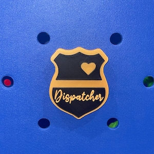 May include: A badge-shaped accessory with a black shield and gold border. The badge features a gold heart and the word "Dispatcher" in a cursive font. The background is a textured blue surface with circular holes.